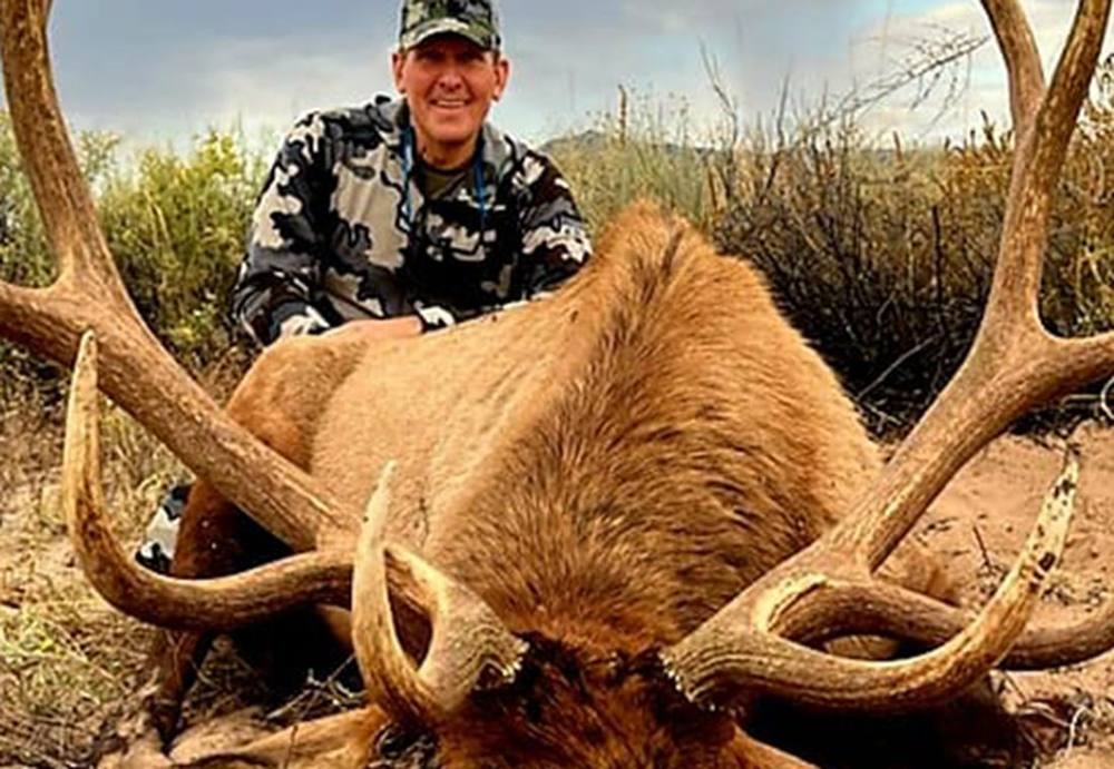 Hunter in camo behind a broad-antlered bull elk in the desert scrub